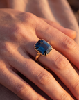 Hand wearing a ring with a blue gemstone against a blurred background