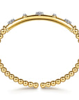 A gold bangle bracelet with a polished curved top band accented by small clear stones, and a beaded lower band made of round gold beads, shown on a white background. side view