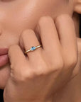 Close-up of a hand wearing a gold ring with a blue gemstone, held near the face.