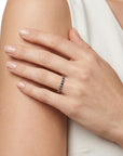 A close-up of a woman's hand resting on her arm, wearing a yellow gold ring with round sapphires. The background is a clean white studio setting.
