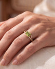 close up of a hand wearing a yellow ring with a green center and diamonds on a neutral background