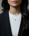 A close-up of a model wearing a delicate gold chain necklace with a small rectangular pendant, styled with a white t-shirt and black blazer.