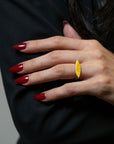 A close-up of a hand with dark red manicured nails wearing a textured yellow gold ring, set against a black top and dark hair.
