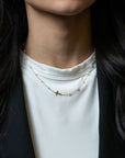 A close-up of a person wearing a delicate gold necklace with a small cross pendant over a white t-shirt and black blazer.