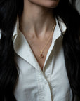 A close-up of a woman wearing a delicate gold chain necklace with a small teardrop-shaped garnet pendant, styled with a white button-down shirt.