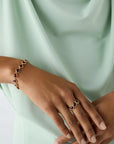 A close-up of a person wearing a gold bracelet and matching ring, each featuring oval red gemstone accents, styled with a mint green draped top.