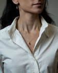 A close-up of a woman wearing a delicate gold chain necklace with a small round pendant and pear shaped garnet drop earrings, paired with a white button-down shirt.