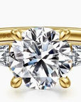 A yellow gold diamond ring with a large round center diamond and two smaller baguette side diamonds, shown on a clean white background.