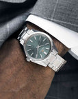 Silver watch with a dark green face on a wrist, wearing gray pants.