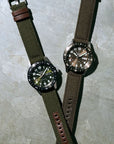 Two watches with green and brown straps on a textured surface