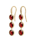 A pair of gold drop earrings featuring three faceted round red gemstones in a textured gold bezel setting, suspended from gold French hooks with a small clear accent stone near the top.