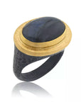 A hand-wrought Lika Behar Pompei statement ring, featuring an east-west oval cabochon labradorite displaying shifting blue and gray flashes, bezel-set in rich 24k yellow gold with a hammered oxidized sterling silver border.
