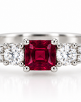 A platinum ring featuring a central square red ruby gemstone flanked by two round brilliant-cut diamonds, with additional small diamonds set along the shoulder, shown on a white background.