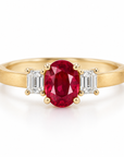 A yellow gold ring featuring an oval-cut red ruby center stone flanked by two emerald-cut clear diamonds, set against a clean white background.
