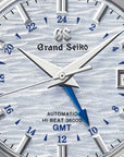 Detailed close-up perspective view of the Grand Seiko SBGJ249 watch face, highlighting the fine, water-ripple dial texture in an icy light blue hue, multi-faceted silver index markers, and the contrasting temper-blue GMT hand.