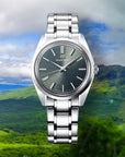 Silver watch with a greenish face on a scenic background of mountains and greenery