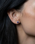A close-up of a person's ear wearing a small round stud earring with a blue center set in a gold-tone bezel, with dark hair framing the side of the face.