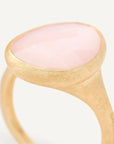 A close-up shot of a gold signet ring featuring a smooth, oval-shaped pale pink stone, set against a clean, light off-white background.