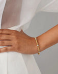 A close-up of a person's hand and wrist wearing a slim gold bangle bracelet with small silver-toned rectangular accents, styled with a white draped garment against a soft gray background.