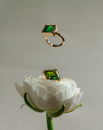 Two gold rings with rectangular green gemstones are shown against a soft gray background, one floating above and one resting on a white flower.