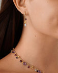 Close-up of a woman wearing a gold gemstone necklace and matching drop earring with small purple and pale blue gemstones