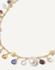 A delicate gold chain necklace adorned with small multicolored gemstones and white freshwater pearls, arranged in a circular shape on a light cream background. close up