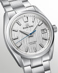 A Grand Seiko stainless steel wristwatch with a textured silver dial, silver-tone metal bracelet, and a distinct blue second hand, shown against a light gray gradient background.
