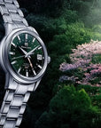 Silver watch with a green dial against a natural background with trees and flowers.