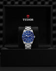 Tudor watch with blue face and silver case on a black display stand against a dark textured background.