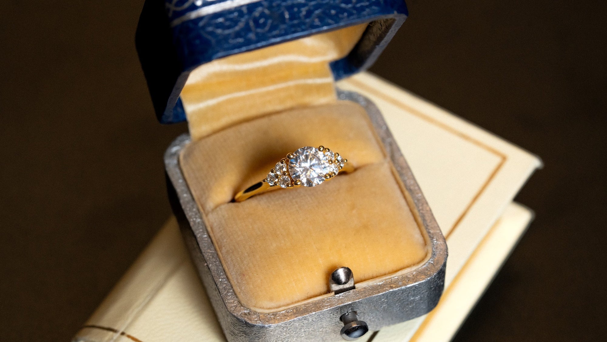 A vintage style wedding ring that features a round center stone and cluster diamonds on the side, in a blue vintage leatherette case