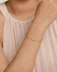 A delicate gold chain bracelet with tiny bead details worn on a woman's wrist, shown against a soft beige pleated top.