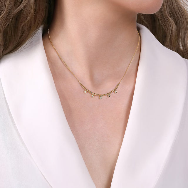 A close-up of a model wearing a delicate gold necklace with small dangling clear stones, styled with a white collared blouse.