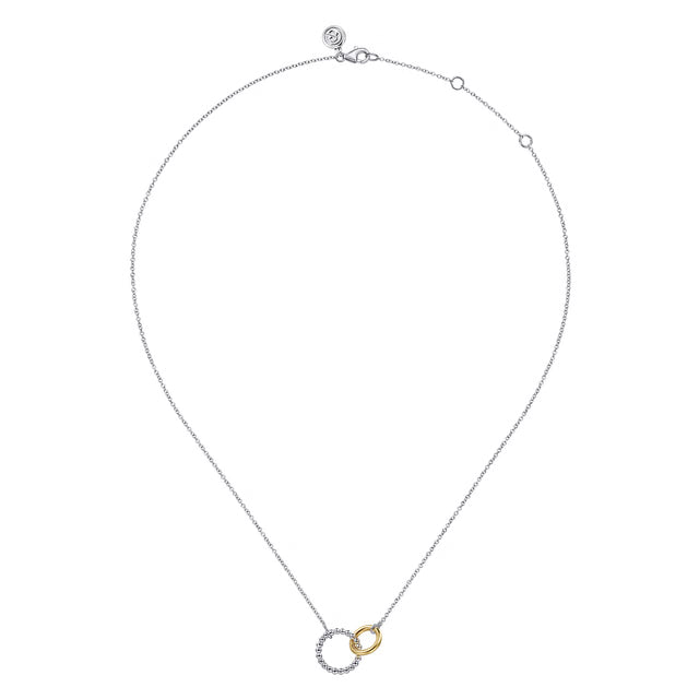 A delicate silver chain necklace featuring a pendant with two interlocking rings, one textured silver and one smooth yellow gold, displayed on a solid white background.