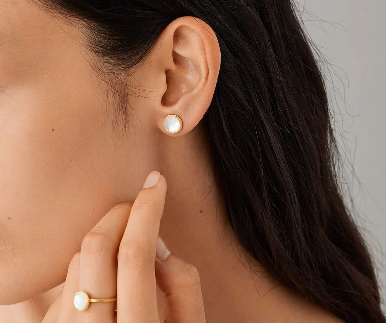 Close-up of a person wearing pearl earrings and a ring, with a neutral background.