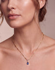 person with curled hair wearing a gold necklace with a bar chain and a blue oval pendant on a neutral background