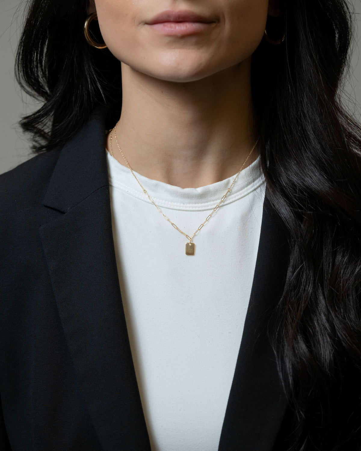 A close-up of a model wearing a delicate gold chain necklace with a small rectangular pendant, styled with a white t-shirt and black blazer.