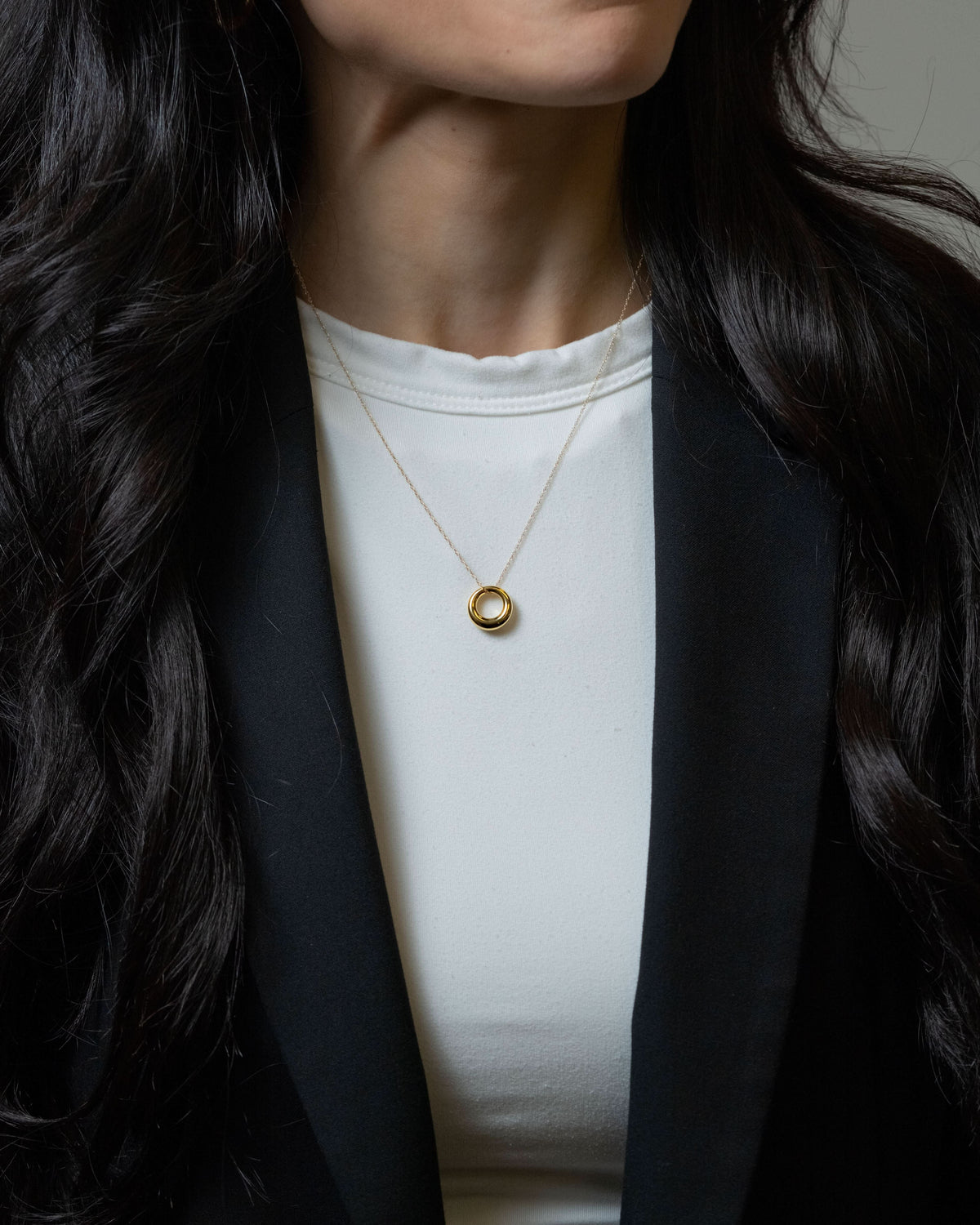 A close-up of a woman wearing a delicate gold chain necklace with a small, polished gold ring pendant, styled over a white top and black blazer.