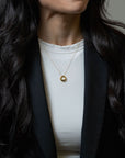 A close-up of a woman wearing a delicate gold chain necklace with a small, polished gold ring pendant, styled over a white top and black blazer.