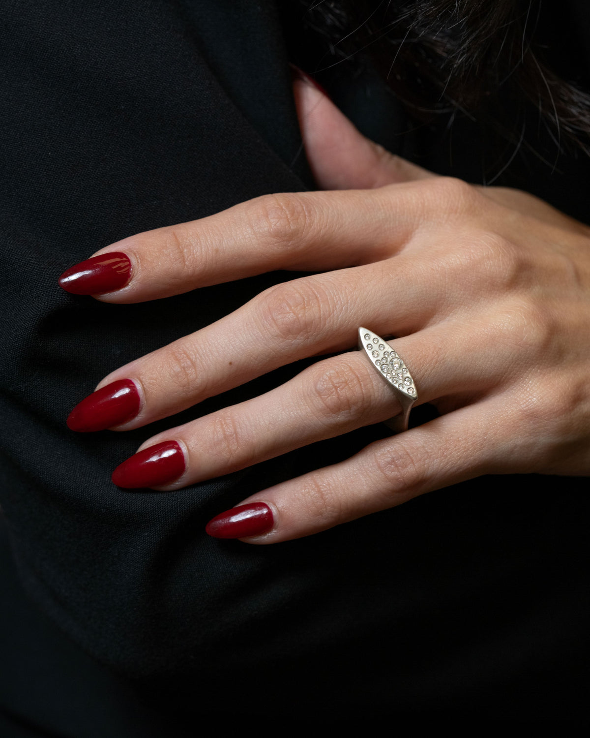 A close-up of a hand with dark red manicured nails wearing a silver marquise-shaped ring set with small clear stones, resting against a black garment background.