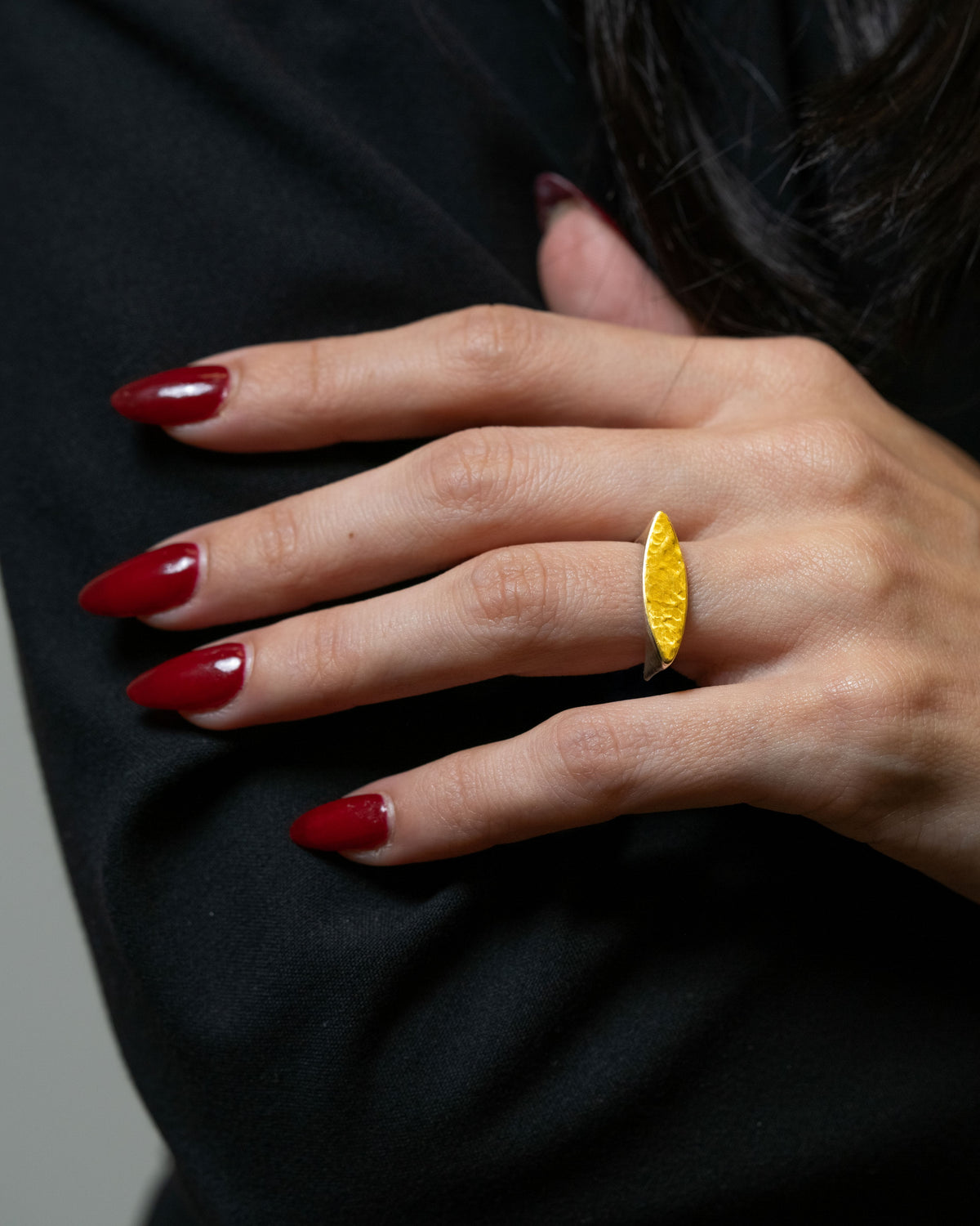A close-up of a hand with dark red manicured nails wearing a textured yellow gold ring, set against a black top and dark hair.