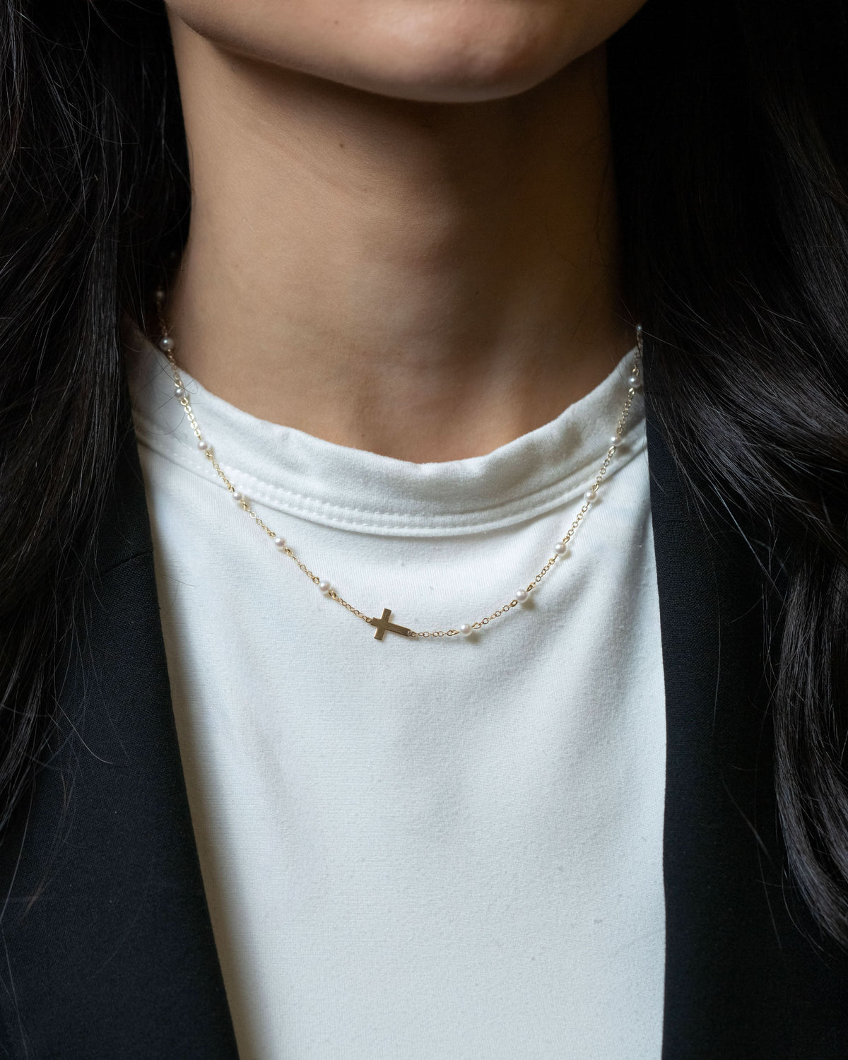 A close-up of a person wearing a delicate gold necklace with a small cross pendant over a white t-shirt and black blazer.