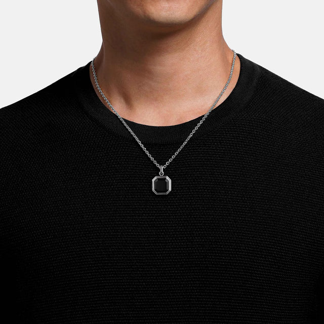 Person wearing a silver necklace with a hexagonal pendant on a plain background