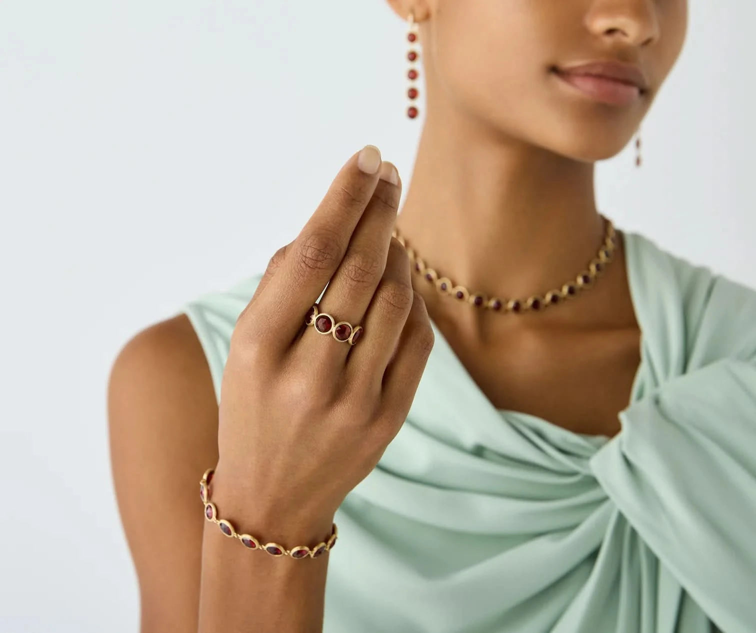 A close-up lifestyle shot of a model wearing gold jewelry, featuring a ring with  red gemstones on her finger, a matching beaded necklace, and a bracelet, set against a clean white background.