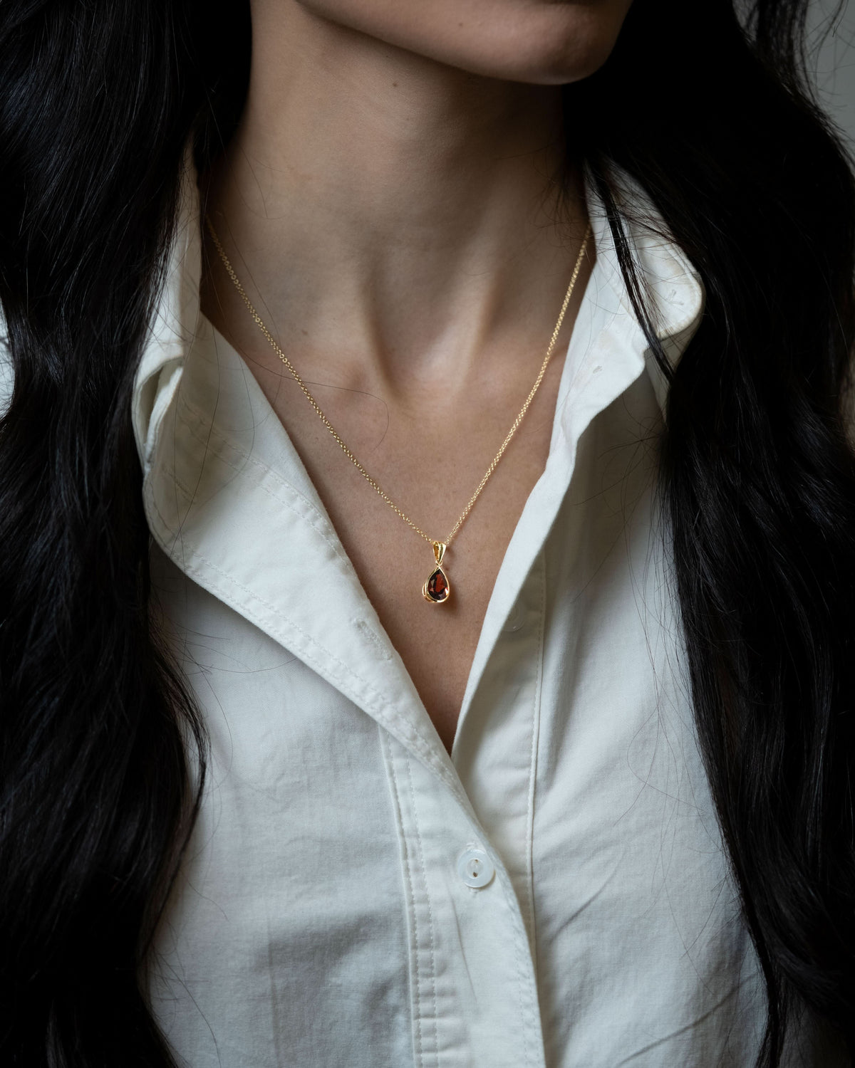 A close-up of a woman wearing a delicate gold chain necklace with a small teardrop-shaped garnet pendant, styled with a white button-down shirt.