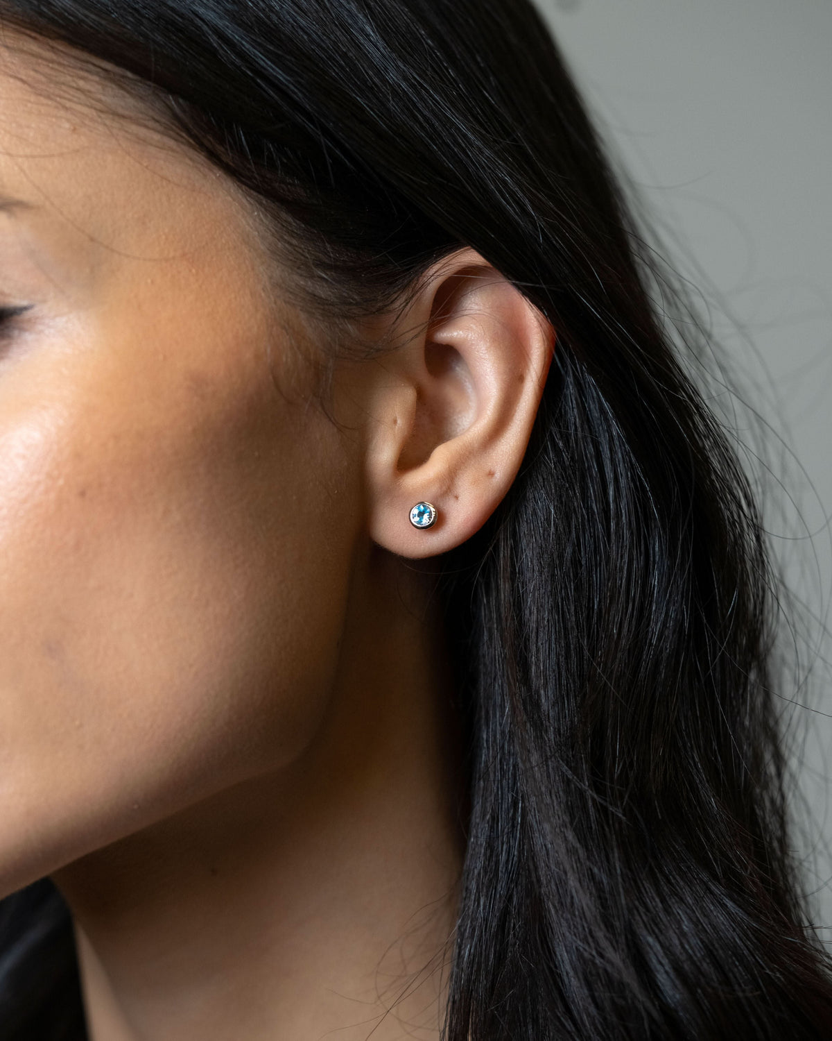 A close-up of a woman's ear wearing a small round stud earring with a blue and white pattern, set against a neutral gray background.