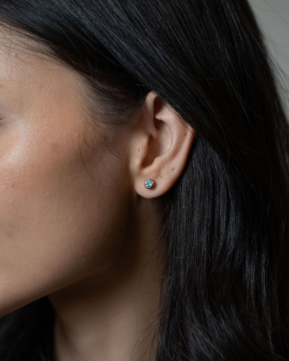 A close-up side profile of a person with dark hair wearing a small round stud earring with a light blue stone and a gold-toned setting.