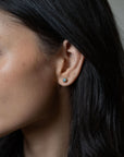 A close-up side profile of a person with dark hair wearing a small round stud earring with a light blue stone and a gold-toned setting.