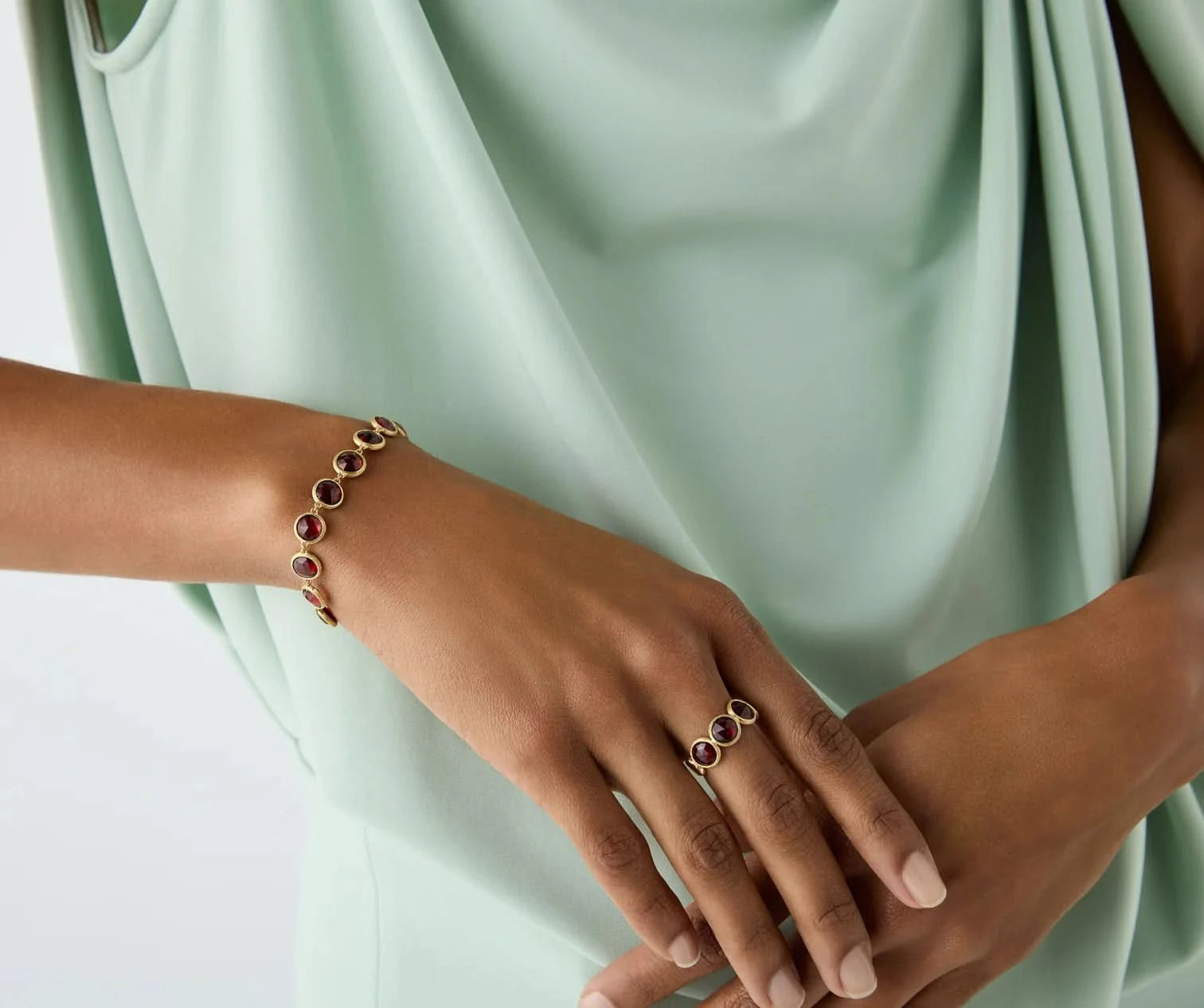 A close-up of a person wearing a gold bracelet and matching ring, each featuring oval red gemstone accents, styled with a mint green draped top.
