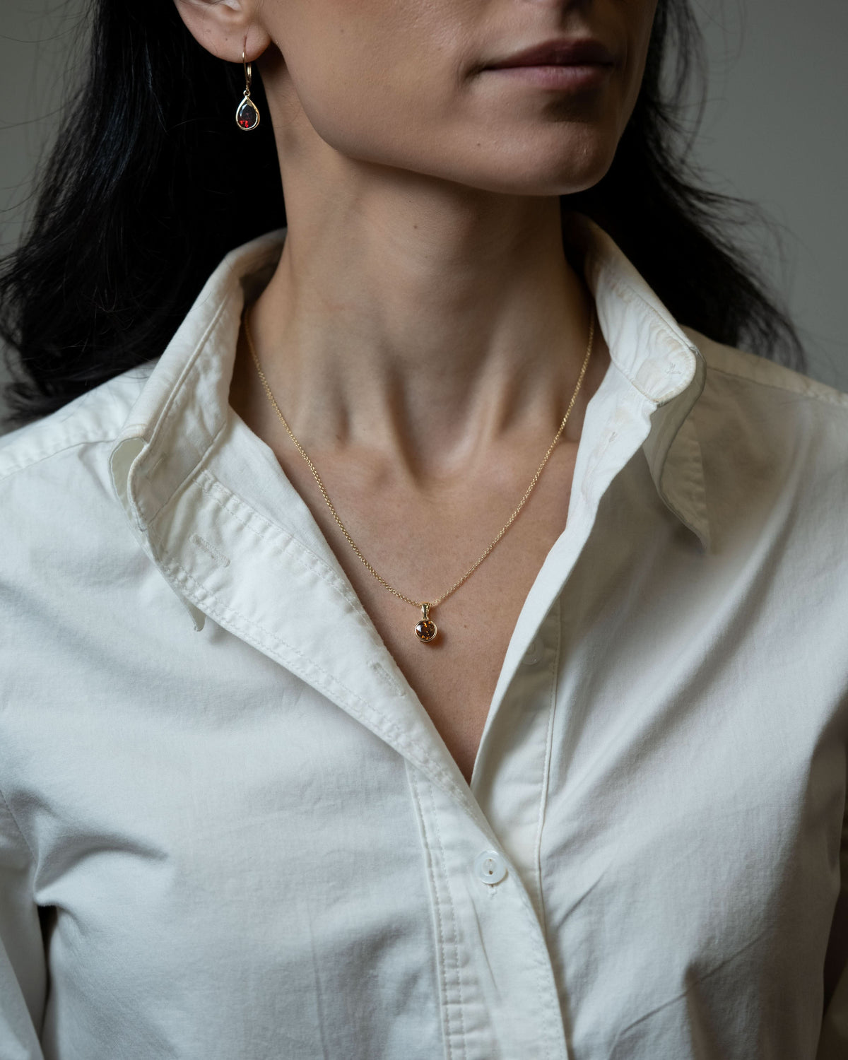 A close-up of a woman wearing a delicate gold chain necklace with a small round pendant and pear shaped garnet drop earrings, paired with a white button-down shirt.