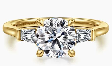 A yellow gold diamond ring with a large round center diamond and two smaller baguette side diamonds, shown on a clean white background.